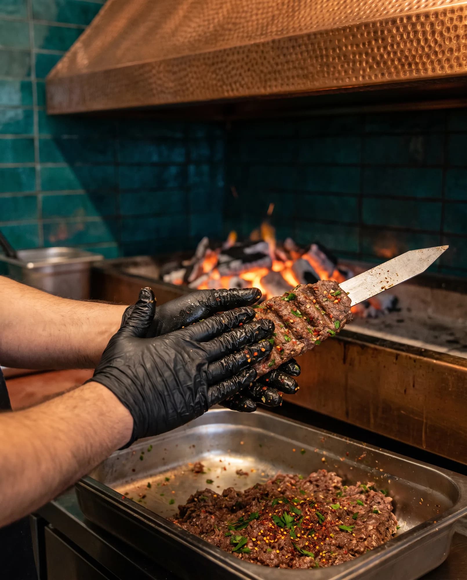 Des mains gantées de noir pressent la viande hachée d'une brochette d'Adana sur une brochette plate, devant la braise